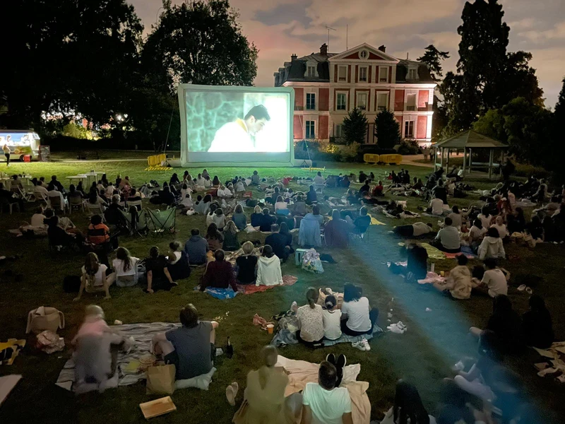 Cinéma en plein air dans un parc devant une demeure, avec écran et public.